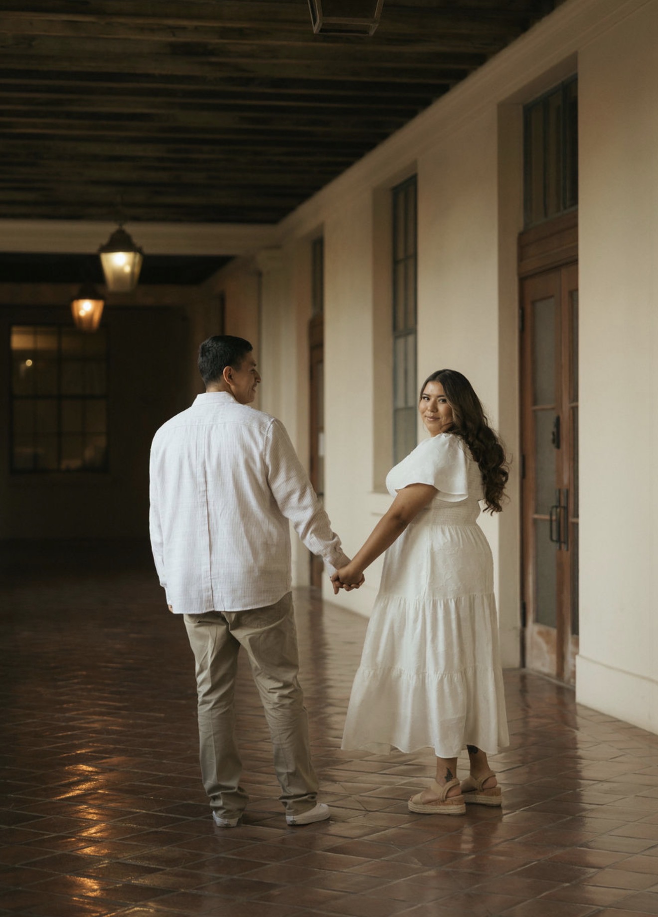 Natalie and Julian holding hands and looking back in a sunlit corridor